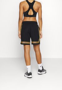 Czarny sportowy biustonosz z wycięciem na plecach, w zestawie z czarnymi szortami z żółto-fioletowymi paskami po bokach oraz szarymi butami sportowymi.