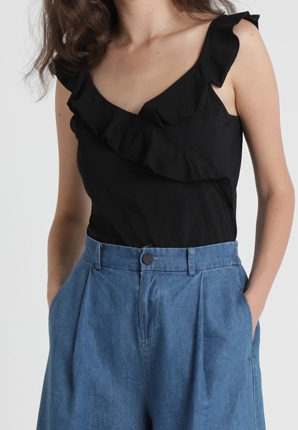 Femme portant un haut noir sans manches avec un col à volants et un pantalon en jean bleu ample taille haute, mains dans les poches.