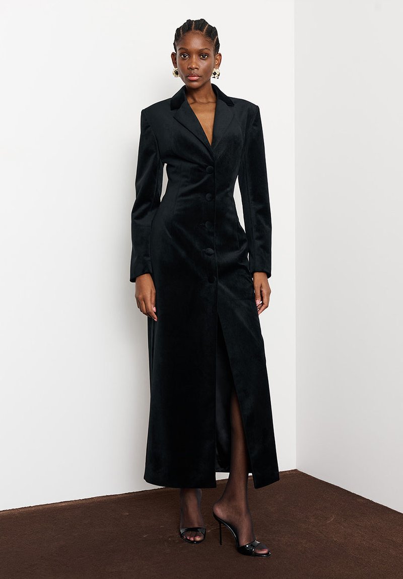 Hajfonatos nő hosszú fekete gombos bársonydresszben és fekete magassarkú szandálban, fehér falnak és barna padlónak támaszkodva.