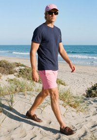Mann mit rosa Kappe, Sonnenbrille, Marinehemd und rosa Shorts, der am sandigen Strand nahe dem Ozean unter klarem Himmel geht.