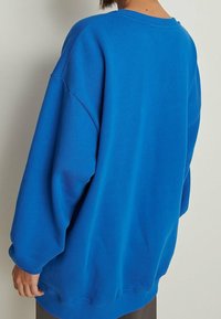 Nadrozměrná modrá mikina s kulatým výstřihem, spadlými rameny a žebrovanými manžetami. Vyrobena z měkké, strukturované látky.