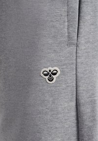 Szara tkanina z małą czarno-białą łatką z logo pszczoły, przyszytą blisko pionowego szwu, prawdopodobnie część ubrania.