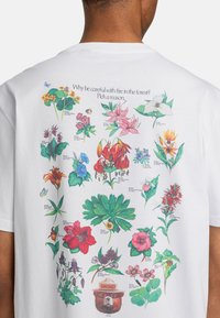 Biela tričko s krátkym rukávom, ktoré na zadnej strane obsahuje farebnú kvetinovú potlač s textom a ilustráciou Smokey Beara v klobúku.