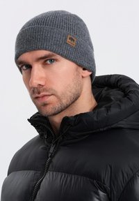 Beanie grigio a maglia con texture a coste e un'etichetta in pelle con il logo di un orso. Indossato con un giubbotto imbottito nero con cappuccio.