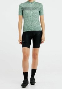 Maillot de cyclisme vert à manches courtes avec un motif à pois et une fermeture éclair à l'avant, associé à un short rembourré noir et des chaussures de cyclisme noires.