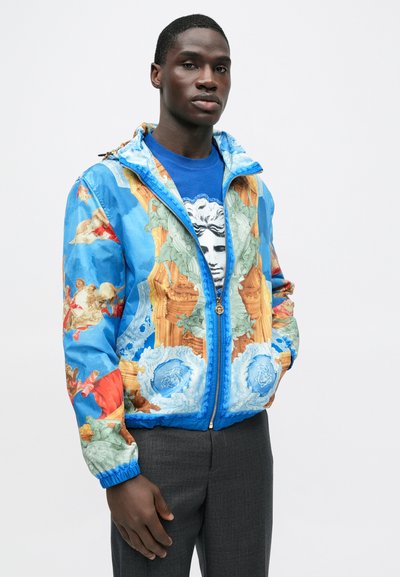 Pisani tiskani suknjič z mešanico modre, oranžne in zelene barve. Ima zadrgo spredaj, elastične manšete in podrobne umetniške vzorce.