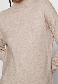 Hellbrauner Strickpullover mit hohem Kragen, lässiger Passform und gerippten Bündchen. Weiche Textur mit einem subtilen melierten Muster.