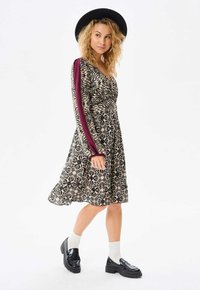 Zwart en crèmekleurig patroonjurk met een V-hals, lange bordeauxrode mouwen en een uitlopende rok. Gecombineerd met zwarte platform instappers en witte sokken.