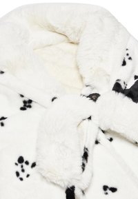 Coperta in finta pelliccia bianca con macchie nere, caratterizzata da una texture morbida. Include un bottone decorativo e bordi ripiegati.