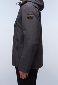 Grå hættetrøjejakke med en lynlåslukket sidelomme og et rundt broderet mærke på venstre ærme. Stoffet ser glat og vandafvisende ud.