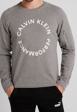 Muž v šedé mikině s dlouhým rukávem Calvin Klein Performance s kruhovým logem a černých kalhotách Puma s logem na stehně.