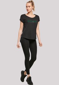 Svart Slytherin t-skjorte med grønn skrift, kombinert med svarte tights og svarte treningssko, med rund hals og avslappet passform.
