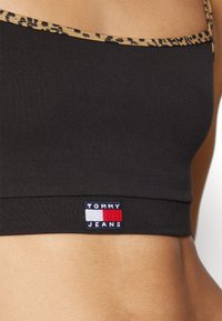 Personne portant un crop top noir avec une bordure à imprimé léopard et un patch logo Tommy Jeans au-dessus de la taille. Seul le torse est visible.