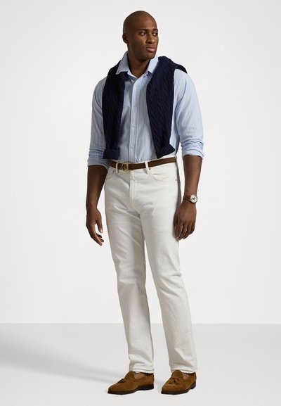 Lys blå stribet skjorte, navy kabelstrik sweater draperet, hvide bukser, brun bælte, brune loafers, iført et armbåndsur.