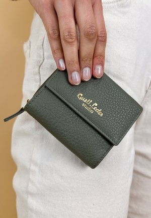 Leather cardholder in dark green with a textured finish. Features a gold logo and a zip closure, held in a hand near light-colored pants.