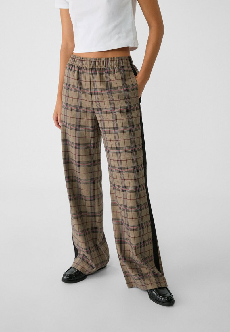 Pantaloni a gamba larga a quadretti in beige con dettagli rossi e neri, dotati di vita elastica e strisce laterali in nero uniforme.