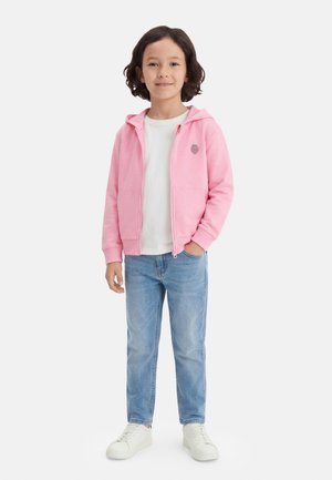 Kind met krullend donker haar draagt een wit T-shirt, roze zip-up hoodie met aardbeienpatch, lichtblauwe spijkerbroek en witte sneakers, staand.