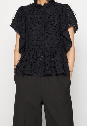 Czarna teksturowana bluzka z krótkim rękawem, falbanką na dole i wzorem wypukłych kropek, zestawiona z czarnymi dopasowanymi spodniami, modelka z rękami w kieszeniach.