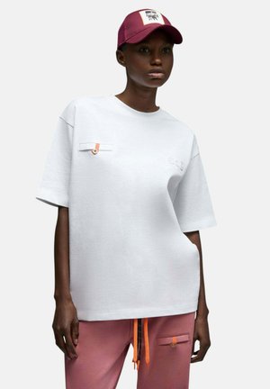 Femme portant un T-shirt blanc oversize avec un détail de poche, un pantalon de jogging rose avec des poches, et une casquette de baseball bordeaux avec un écusson logo.