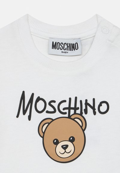 Λευκό βρεφικό μπλουζάκι με μαύρο κείμενο "MOSCHINO" και ένα χαμογελαστό καφέ αρκουδάκι κάτω από το κείμενο. Η ετικέτα στο γιακά γράφει "MOSCHINO BABY."
