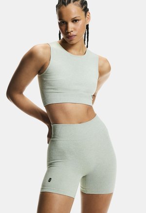 Atlétikus nő, kötött copffal, világosszürke bordázott crop topot és magas derekú rövidnadrágot visel, kezeit csípőjén tartva fehér háttér előtt áll.