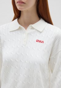 Maglione bianco a trecce con colletto, caratterizzato da ricamo rosso "amour" sul petto e un motivo testurizzato su tutta la stoffa.
