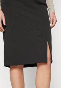 Jupe noire longueur genou avec fente latérale portée par une personne à la peau claire et un haut beige à manches longues, mains reposant sur les côtés.