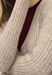 Beige pletený kardigan s ribem a hlubokým V-výstřihem, přes který je navlečen bordový top, ukazující měkkou texturu a teplo.