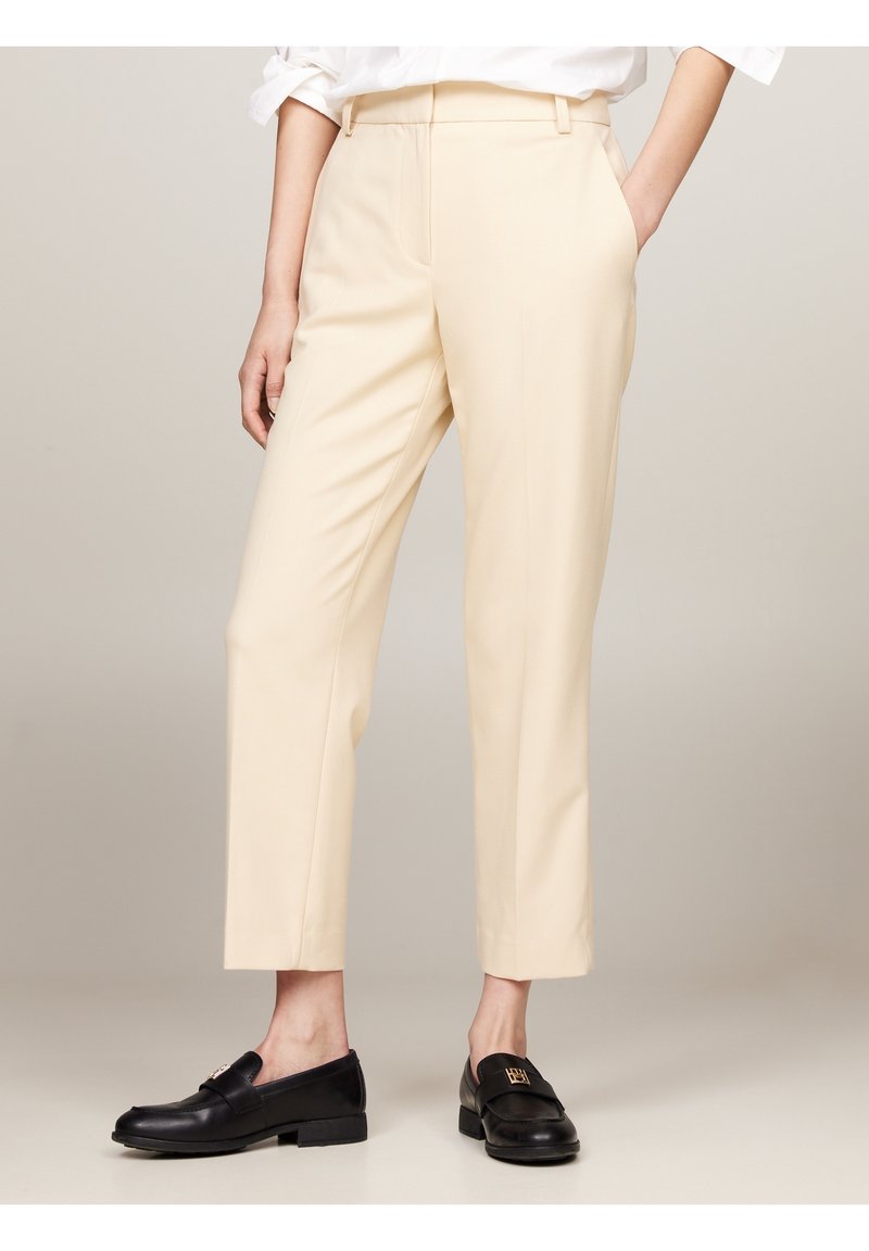 Pantalon beige sur mesure longueur cheville porté avec des mocassins noirs en cuir ornés de boucles dorées, associé à une chemise blanche rentrée.