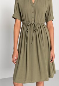 Vestido verde oliva con rayas verticales, mangas cortas enrolladas, botonera y cinturilla ajustable. Textura de tela suave y fluida.