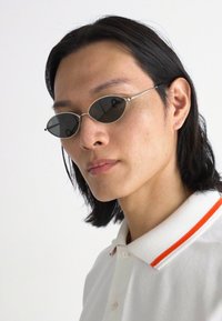 Gafas de sol ovaladas con lentes oscuros, montura delgada de color plateado y un detalle sutil. El modelo lleva una camiseta polo blanca con una raya naranja en el cuello.