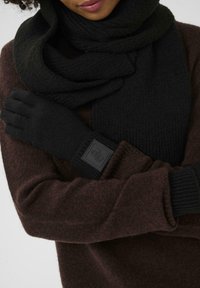 Schwarze Strickhandschuhe mit einer strukturierten Oberfläche, ausgestattet mit einem rechteckigen schwarzen Logo-Patch, kombiniert mit einem dunkelbraunen Pullover und passendem Schal.