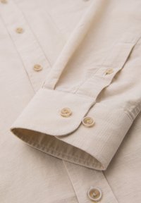 Poignet de chemise beige à manches longues avec deux boutons, patte de boutonnage visible et tissu texturé.