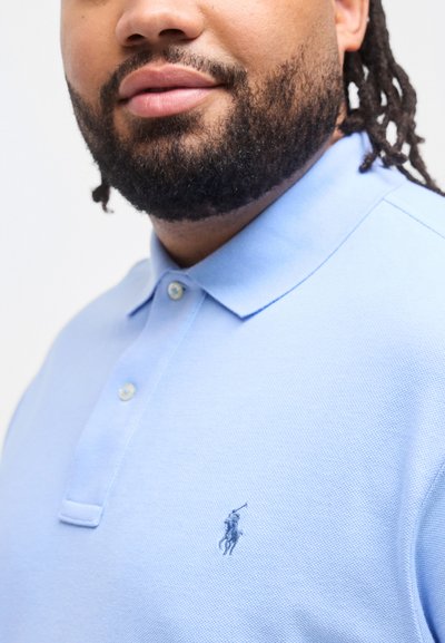 Άνδρας με μούσι και κοτσίδες, φορώντας ανοιχτό μπλε πόλο πουκάμισο με μικρό κεντημένο λογότυπο αλόγου και αναβάτη στο στήθος.