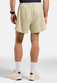 Beige Laufshorts mit elastischem Bund, darüber trägt man hellgrüne Kompressionsshorts. Strukturierter Stoff, lockerer Schnitt, über dem Knie.
