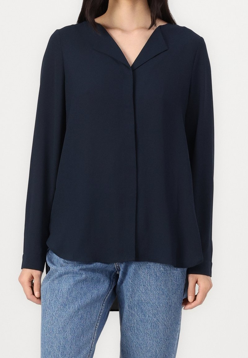 Blusa a maniche lunghe blu navy con scollo a V, tessuto morbido, chiusura frontale con bottoni e vestibilità comoda, abbinata a jeans in denim azzurro chiaro.