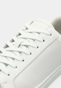 Valge nahast tenniseking valgete paeltega, lähivõte, kus on nähtav õmbluse detail ja osa tallast heledal taustal.