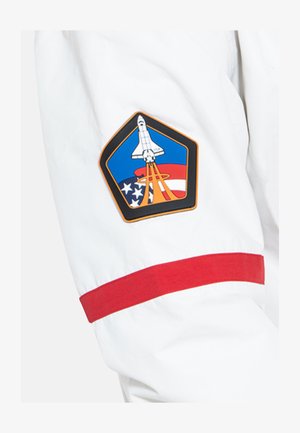 Bílá bunda s červeným lemem na rukávu a vyšívanou nášivkou zobrazující raketoplán, americké vlajky a kruhové ozdoby v modré a červené barvě.
