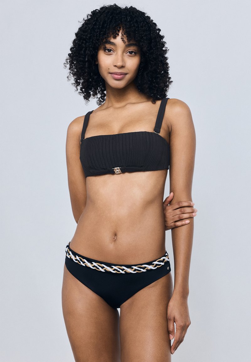 Γυναίκα με σγουρά μαλλιά που φοράει μαύρο μπικίνι με λεπτομέρεια πλεγμένης ζώνης, στέκεται με το ένα χέρι σταυρωμένο πάνω στη μέση της.