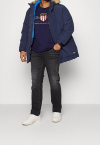 Marinblå parka med pälsfodrad huva, blå insida, bärs över en marinblå grafisk t-shirt, tillsammans med mörka jeans och vita sneakers.