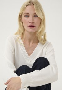 Baltas V-veida krekls ar ribinātu kakla izgriezumu un aprocēm, ar mīkstu tekstūru. Modelis sēž tums zilos džinsos, rokas aptvērušas ceļus.