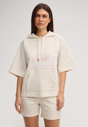 Donna che indossa una felpa beige a maniche corte con cappuccio e motivo a fantasia con il logo "JOOP!" e pantaloncini abbinati, in piedi davanti a uno sfondo chiaro e uniforme.