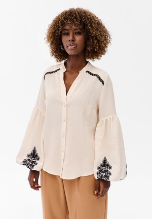 Kobieta o kręconych włosach, ubrana w kremową bluzkę na guziki z czarnymi koronkowymi detalami i balonowymi rękawami, w połączeniu z beżowymi, szerokimi spodniami.