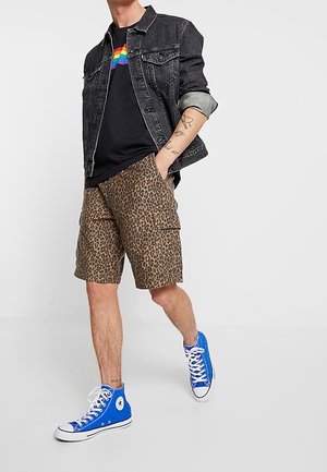 Veste en jean noire aux manches retroussées, t-shirt noir avec un motif arc-en-ciel, short à imprimé léopard et baskets montantes bleues.