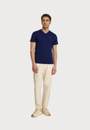 Tânăr stând, purtând un tricou bleumarin cu guler în V și logo roșu, pantaloni bej și adidași bej deschis pe un fundal simplu.