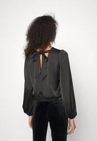 Donna con capelli ricci che indossa una blusa nera di raso a maniche lunghe legata dietro e pantaloni di velluto neri, vista di spalle.