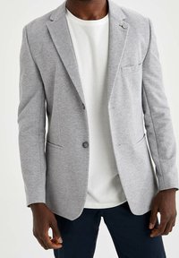 Hellgrauer Blazer aus strukturiertem Stoff, mit einer einzelnen Knopfleiste, angesteppten Revers und einer Brusttasche. Wird über einem weißen T-Shirt getragen.