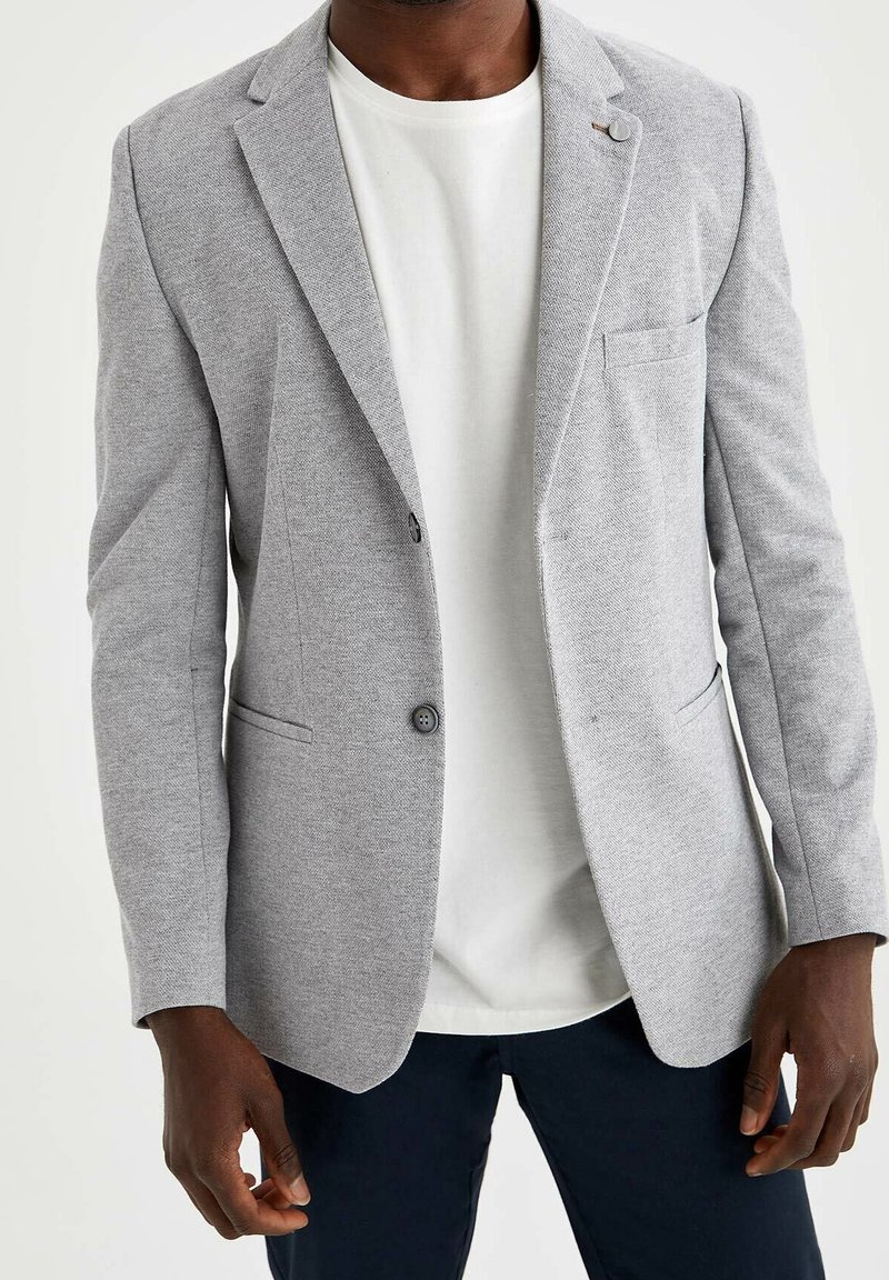 Hellgrauer Blazer aus strukturiertem Stoff, mit einer einzelnen Knopfleiste, angesteppten Revers und einer Brusttasche. Wird über einem weißen T-Shirt getragen.