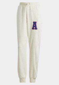 Krämfärgade sweatpants med elastiska muddar, dragsko i midjan, vertikal söm på ett ben och ett stort blått och rött bokstavs-"A"-märke.