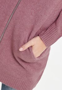 Strickjacke in staubigem Rosa, mit einem Reißverschluss vorne und gerippten Bündchen. Seitentasche sichtbar, der Stoff hat eine weiche, strukturierte Oberfläche.